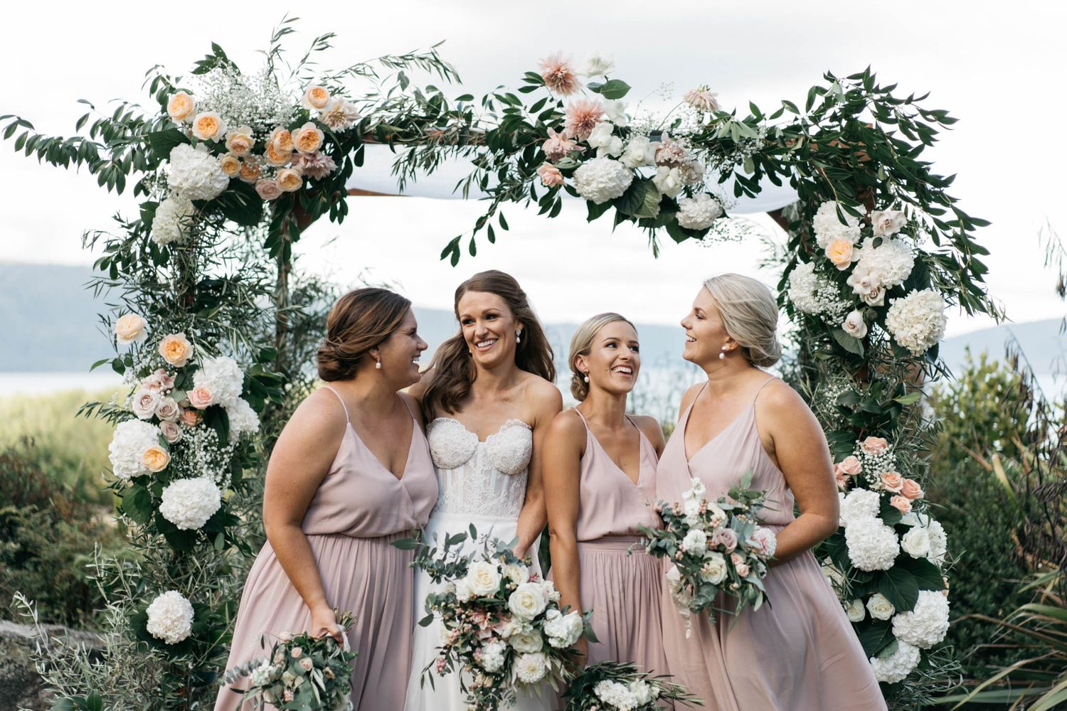 Bridesmaids wearing dusty mauve dresses share a joyful candid moment together in an outdoor garden setting.