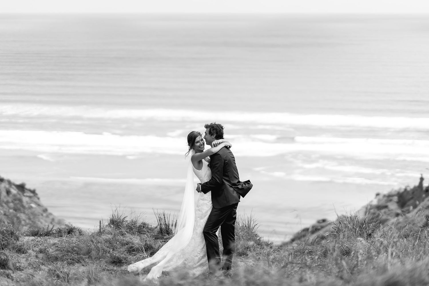 Romantic couple embraces on coastal cliffs overlooking a misty ocean in black and white photography.