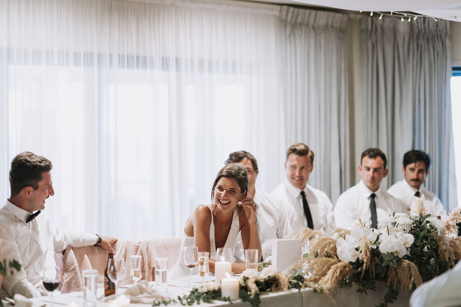 Panoramic view of an elegant white wedding reception setup with long tables and ethereal curtains in natural light.