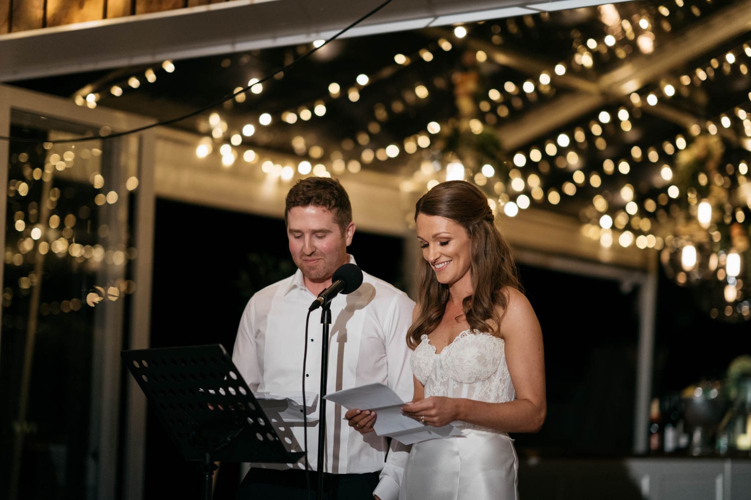 A couple reading speeches outdoors in the evening under glowing string lights at a wedding reception.