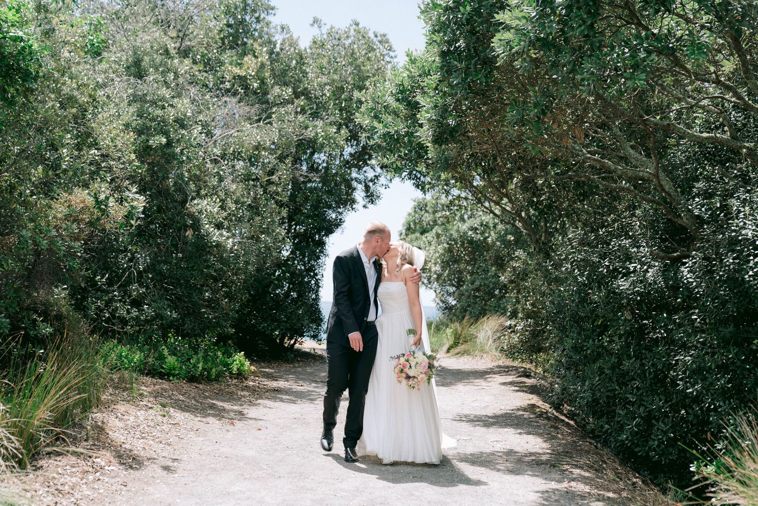 A couple in wedding attire stands on a sunlit path surrounded by lush green foliage.