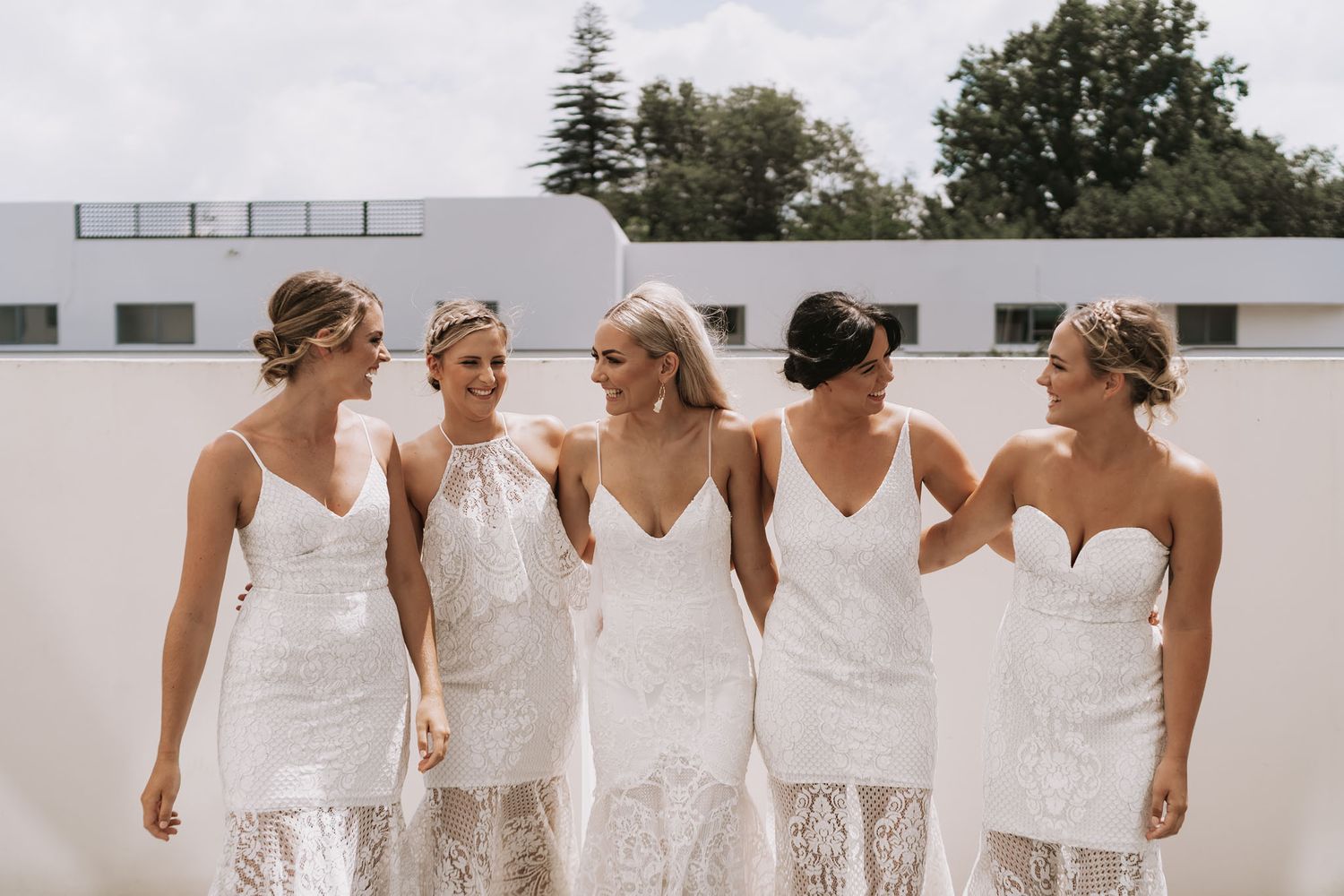 Group of bridesmaids in matching white lace dresses walking together on a sunny day.
