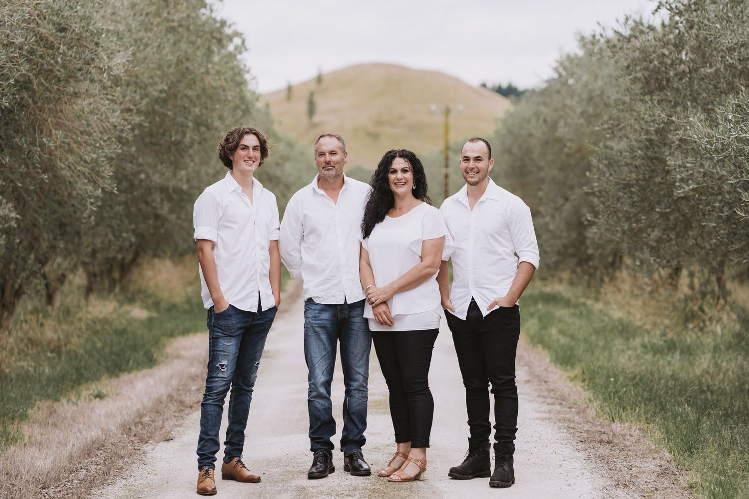 Four people wearing white shirts pose on a dirt road lined with olive trees and rolling hills.