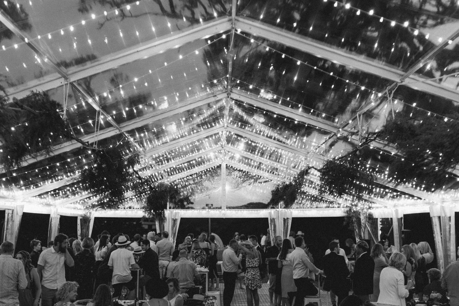 A tented outdoor reception venue illuminated with string lights and filled with wedding guests dancing at night.