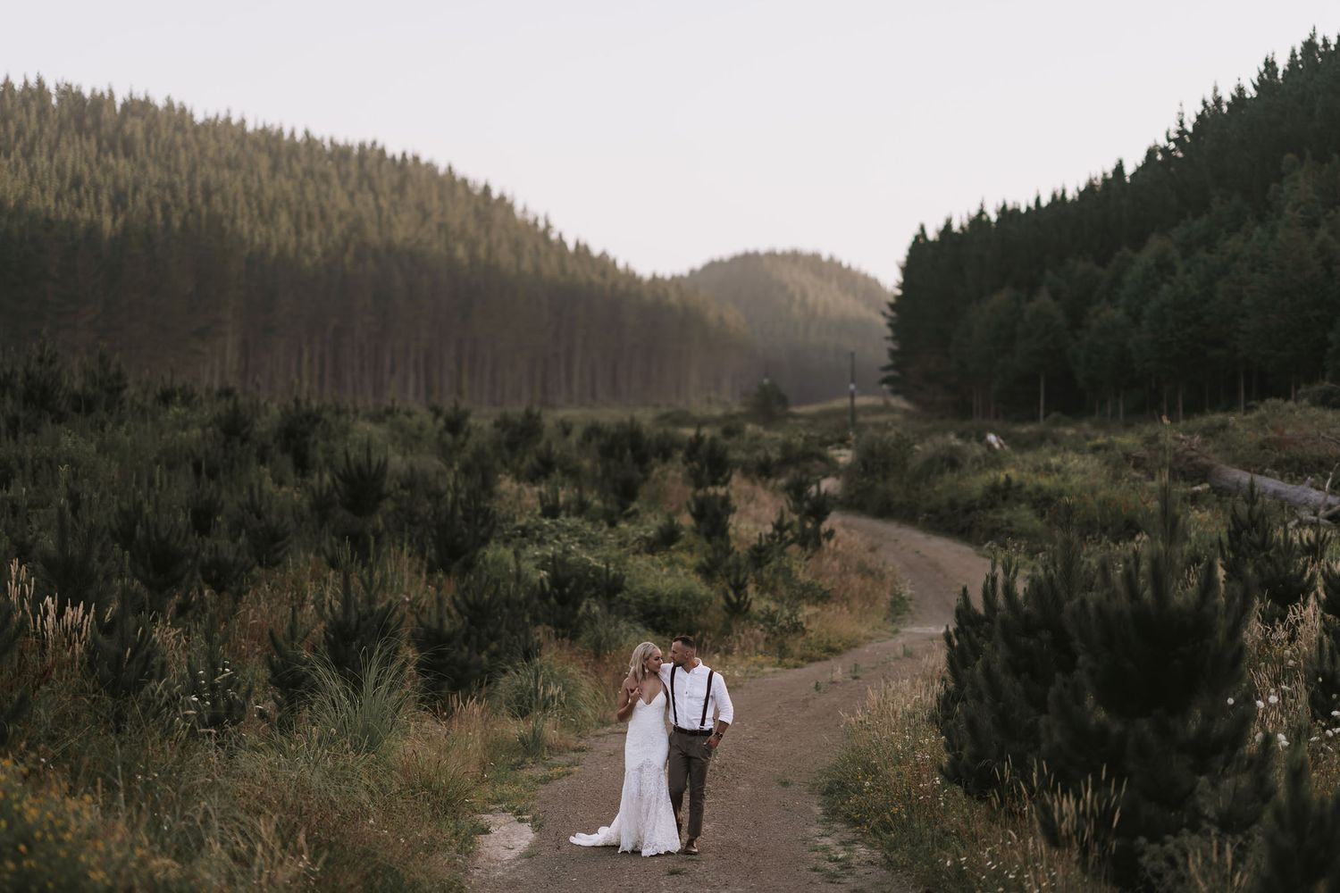 A couple walks down a winding dirt road surrounded by mountainous forest terrain at dusk.