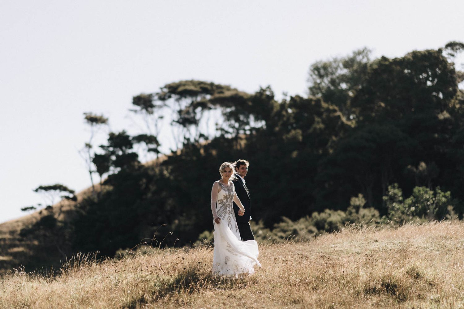 A couple stands together on a windswept hilltop with golden grass and silhouetted trees in the background at sunset.