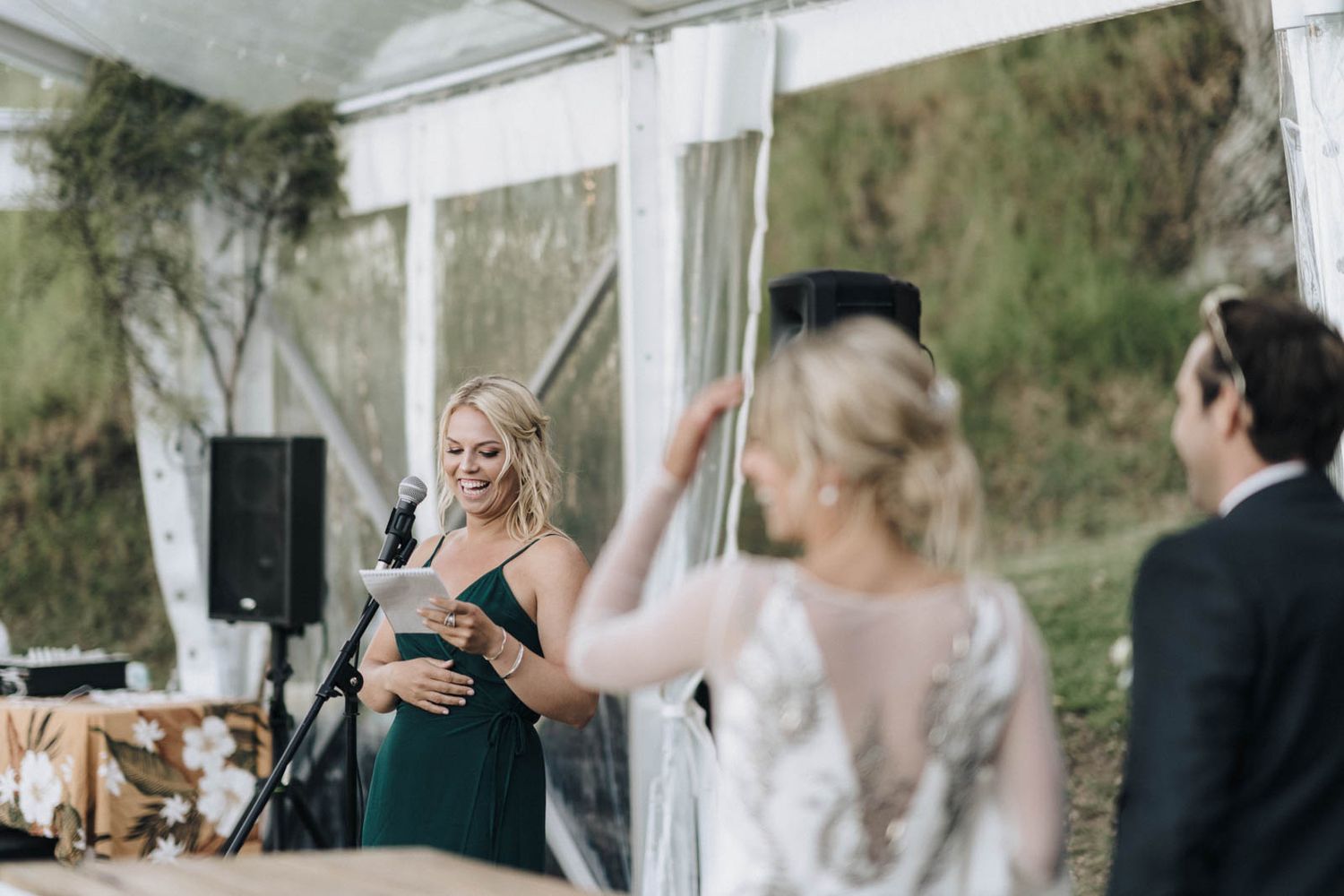 Wedding reception speech under white tent with speaker in emerald green dress and guests listening at outdoor venue.