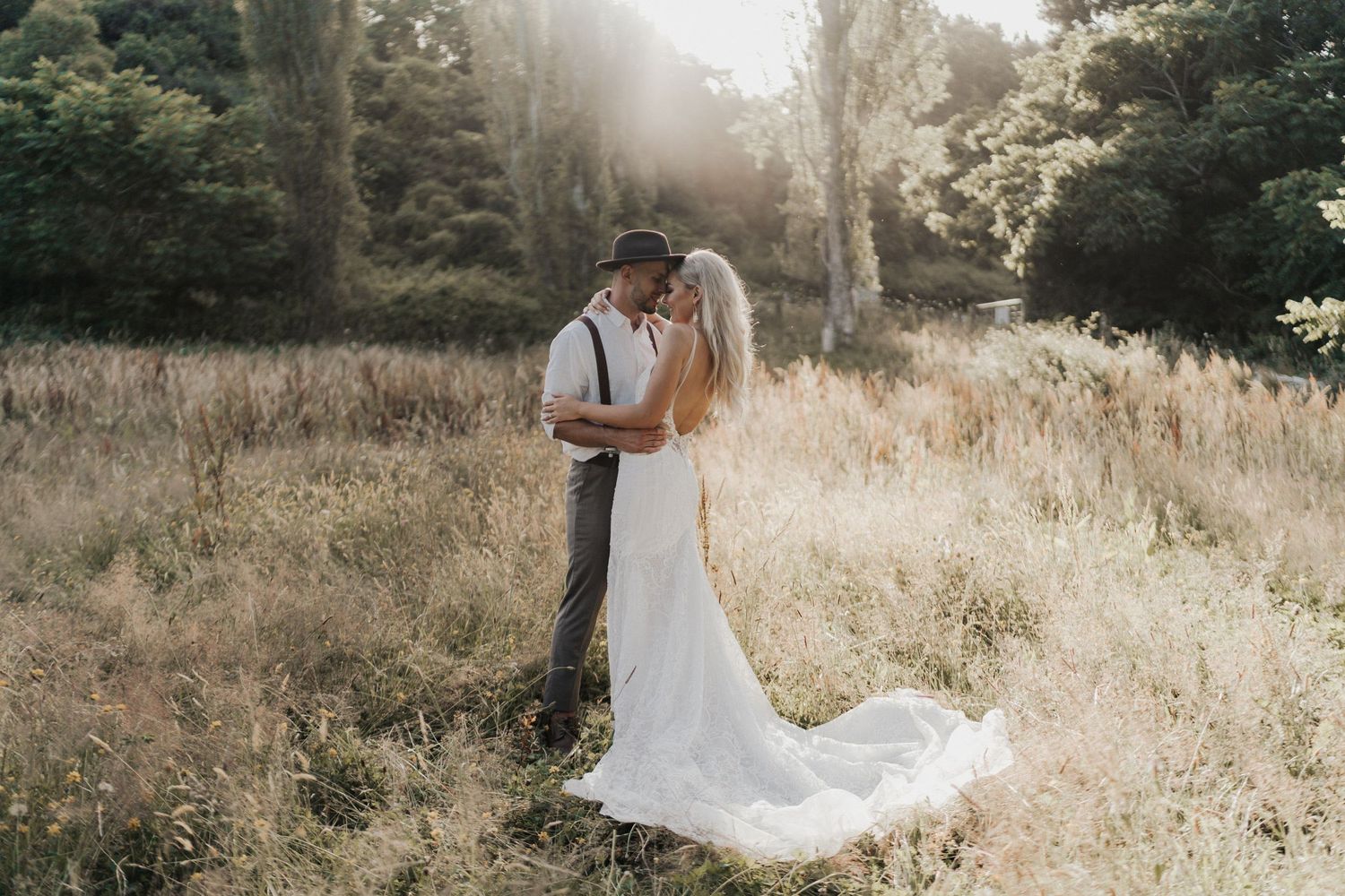 A couple shares an intimate moment in golden sunlit field surrounded by trees.