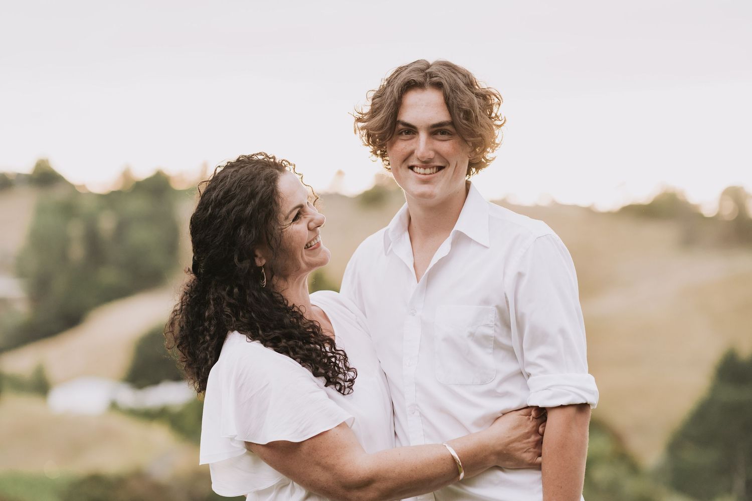 A tender outdoor moment between a couple in white shirts against a dreamy natural backdrop at sunset.