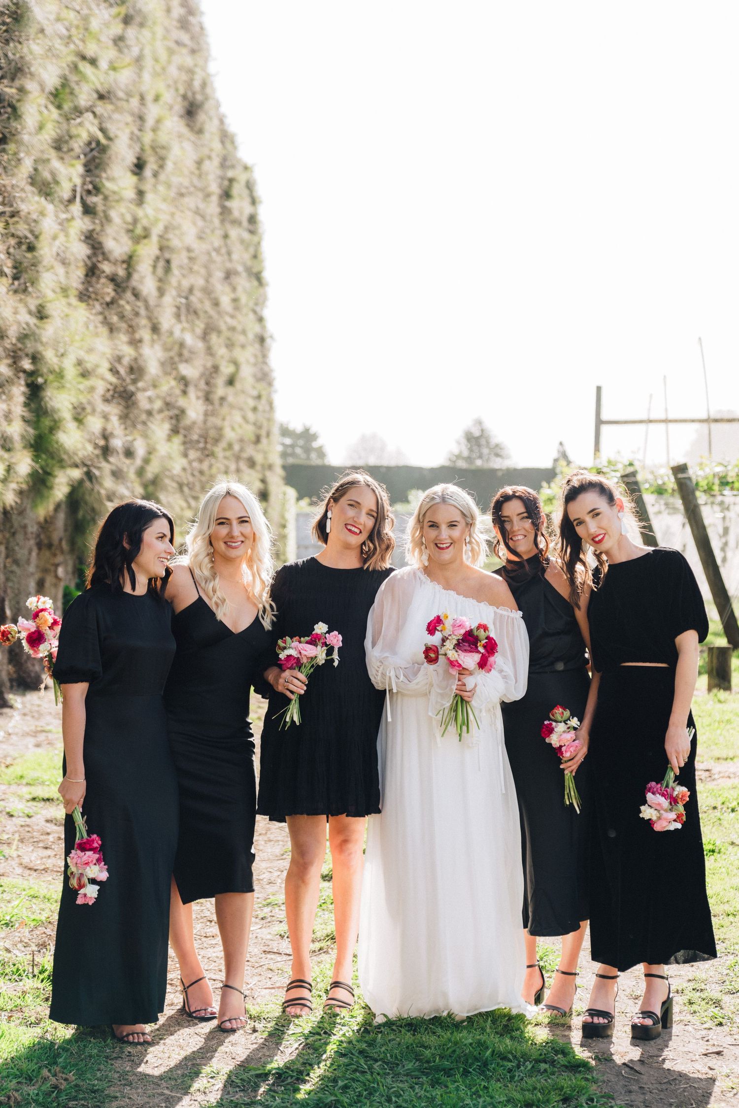 A bridal party in black dresses and white gown pose together outdoors holding pink bouquets.