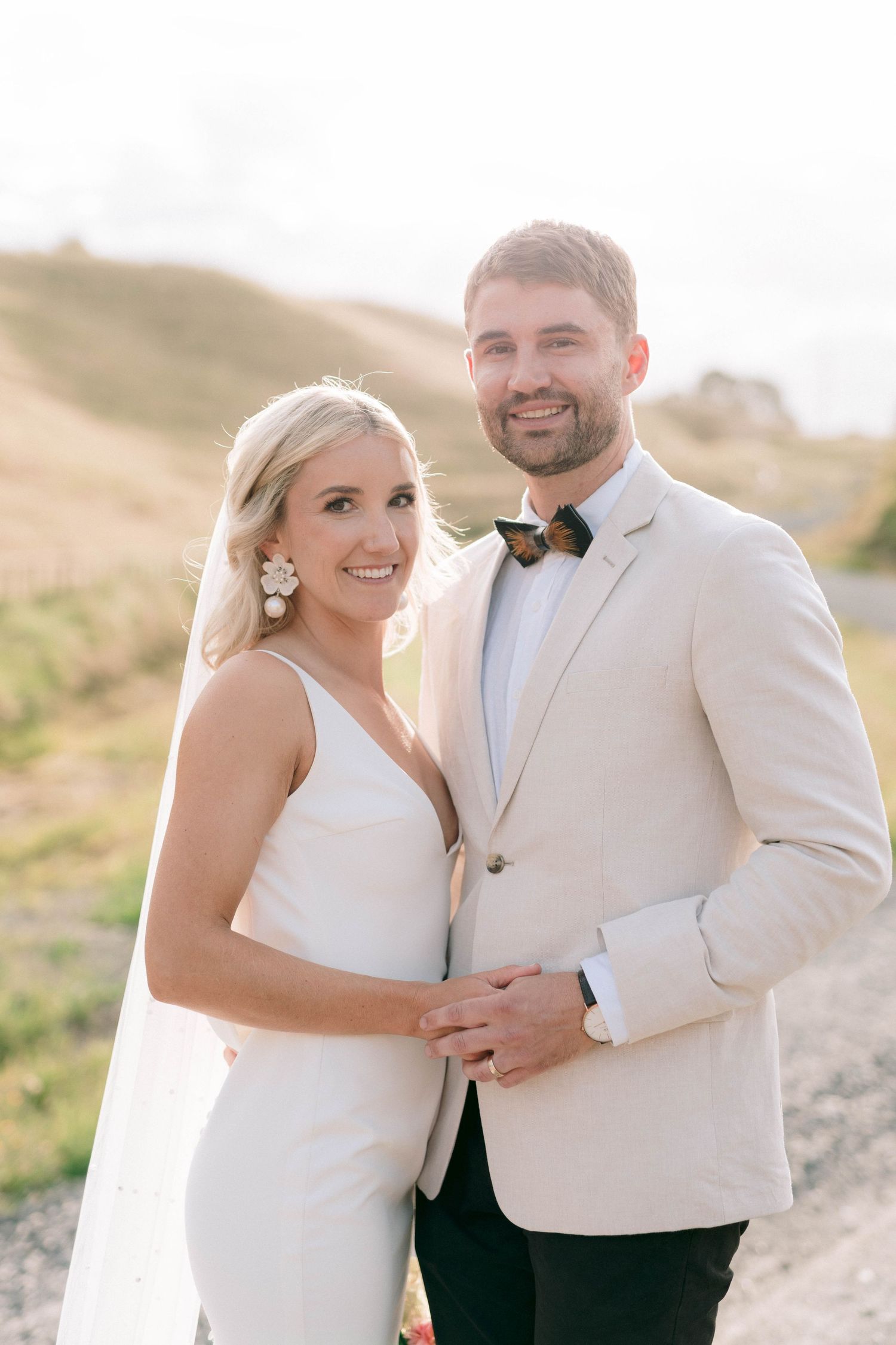 Newlyweds pose together outdoors wearing cream and white formal wedding attire.