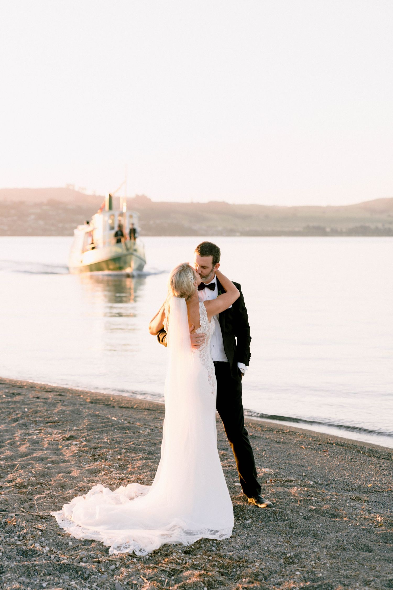 A romantic couple embraces on a beach at sunset with a boat visible in the water behind them.