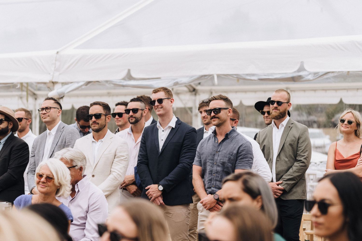 Wedding guests in formal attire watching ceremony under white canopy.