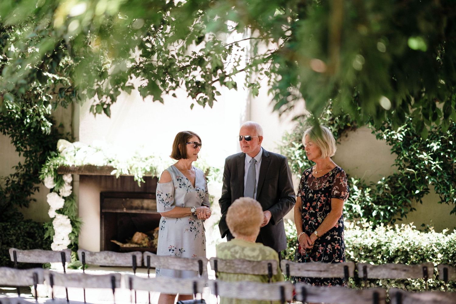 People gather for conversation in a sunlit garden setting with white floral decorations and greenery.