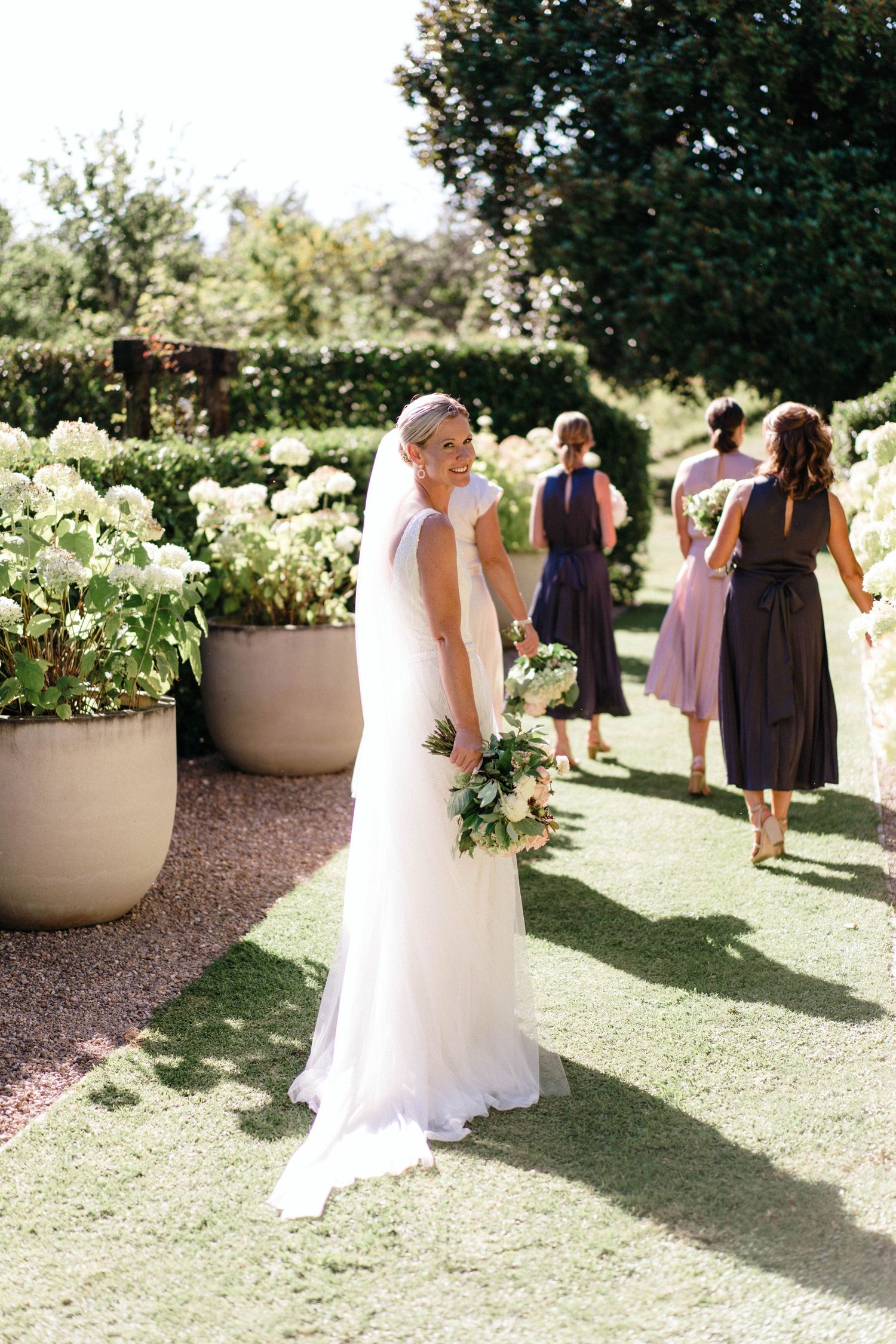 A white wedding dress trails behind figures walking through a sunny garden with decorative planters.