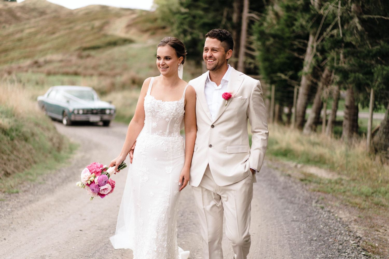 A couple walks down a rural gravel road on their wedding day wearing a white dress and light beige suit.