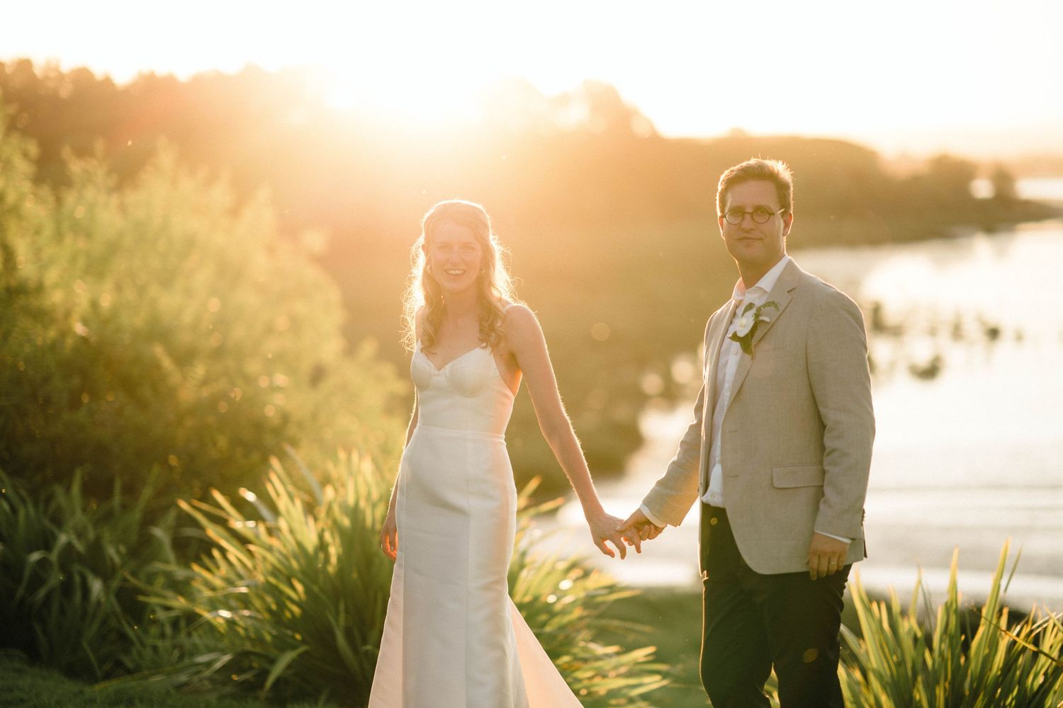 Newlyweds pose together at sunset near waterfront with golden light.