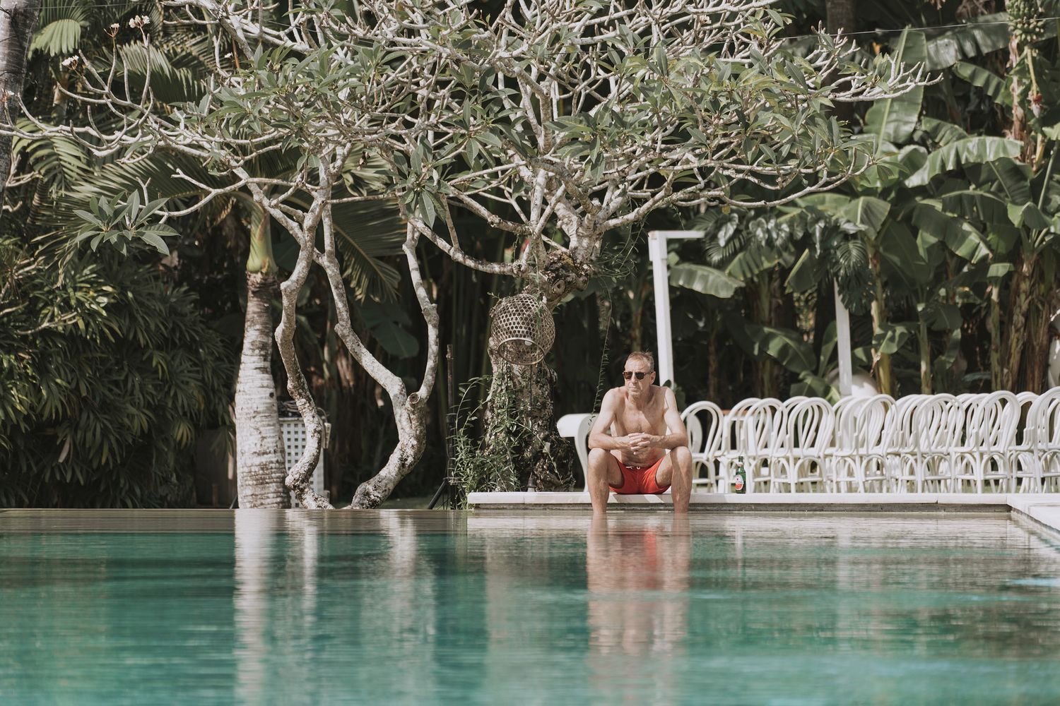 Person relaxing at the edge of a luxurious infinity pool surrounded by tropical trees and white railing.