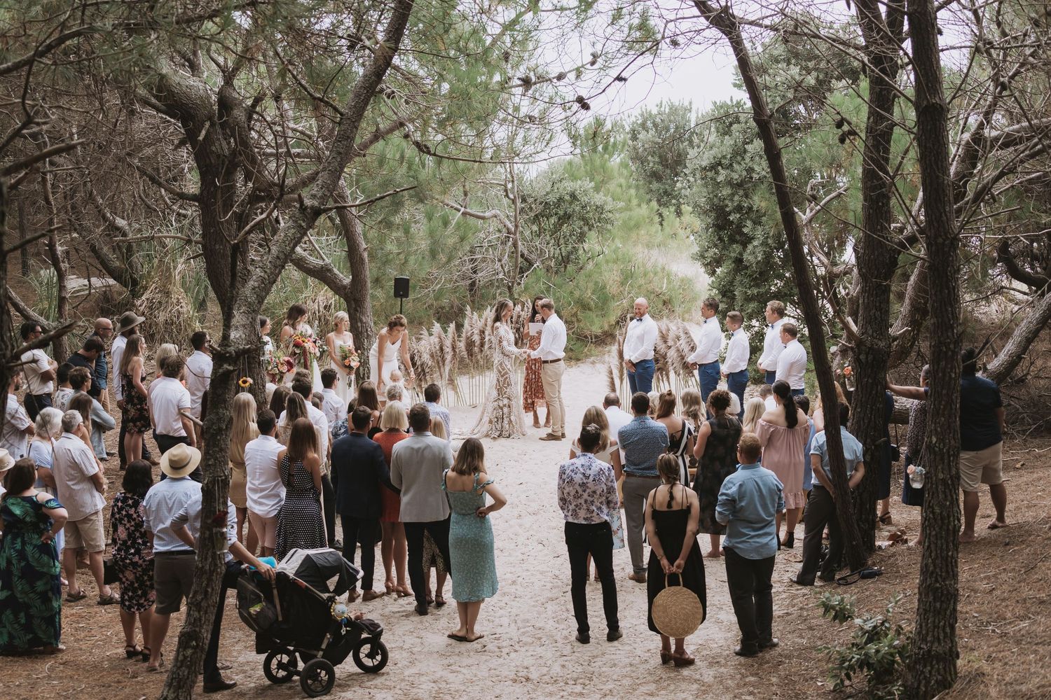 A group of people gathered for an outdoor wedding ceremony on a wooded path surrounded by trees.