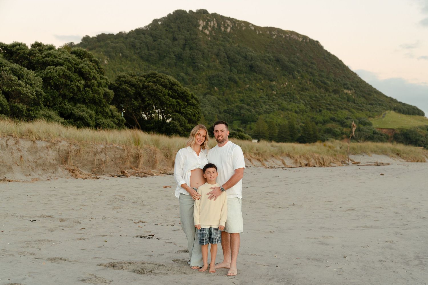 Family of three posing on sandy beach with lush green mountain backdrop at dusk. Photographer Bryony Alexander Newfound.