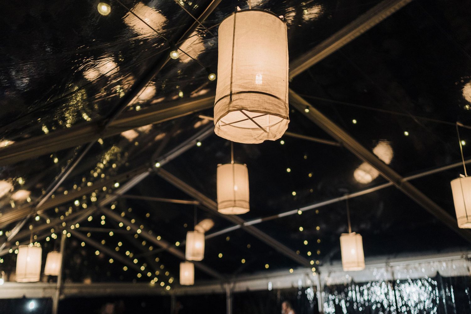 Paper lanterns and string lights create an intimate ambiance under a dark wooden ceiling at an evening venue.