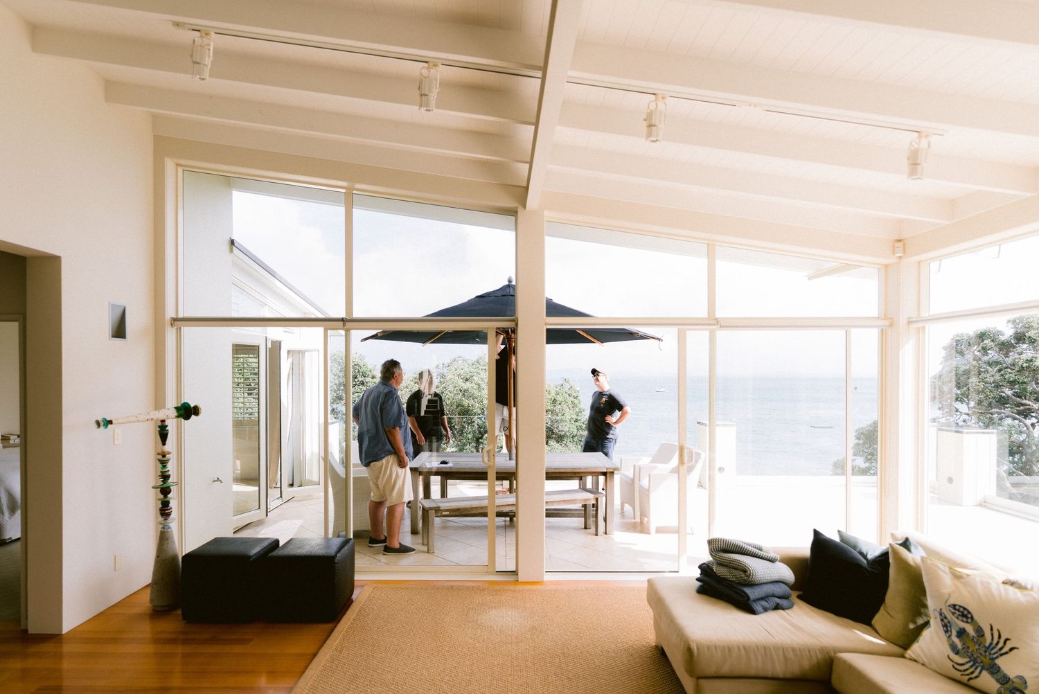 Modern sunlit living room with glass walls overlooking ocean features cozy seating and a dining area.