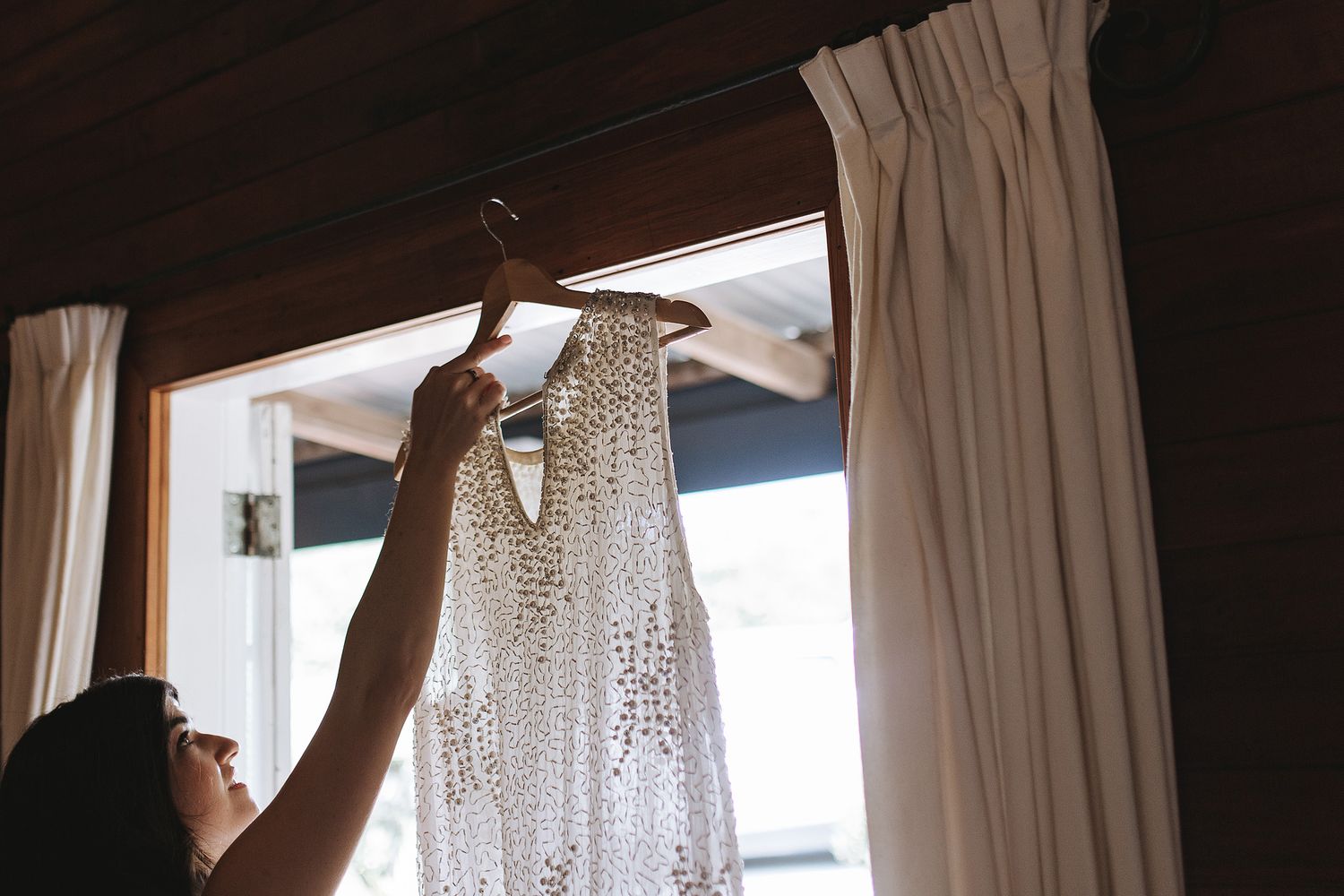 A silhouetted figure holds up a delicate lace wedding dress against natural window light in a room.