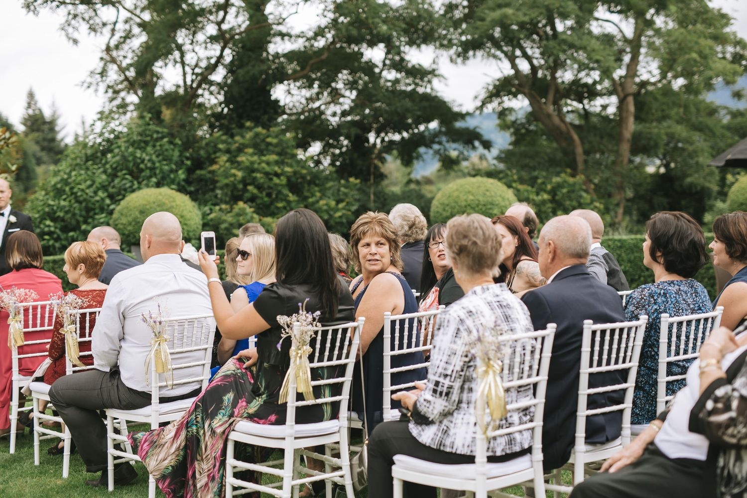 Panoramic view of wedding guests sitting in white chairs during an outdoor ceremony at a scenic venue.