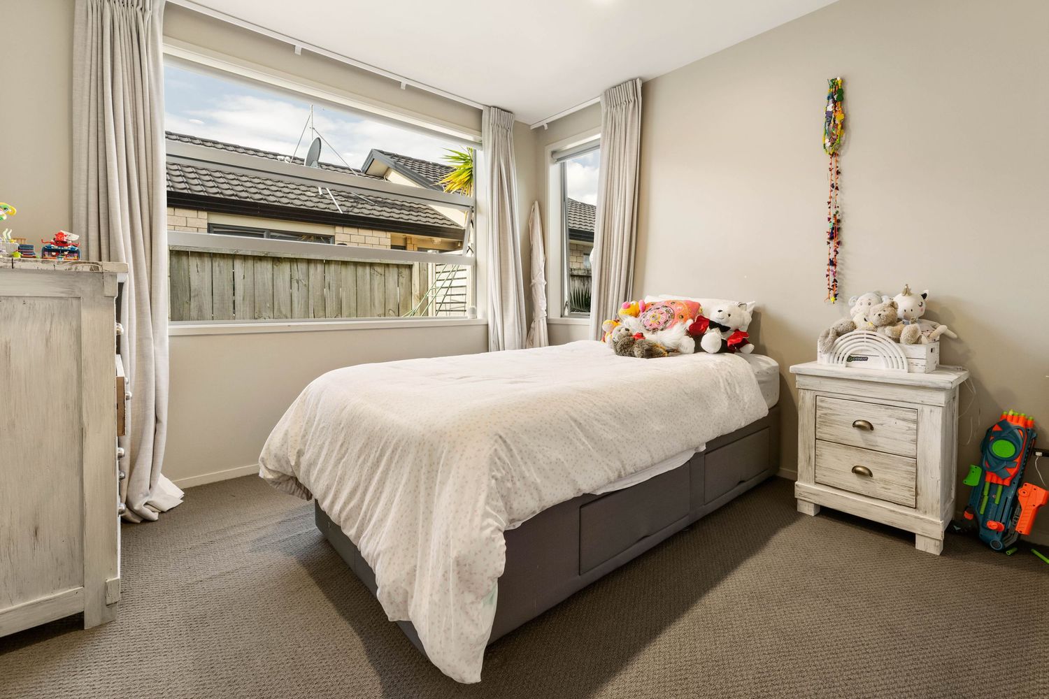 Cozy bedroom with gray carpet featuring a white comforter, bedside table, and curtained window.