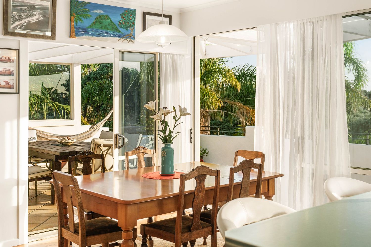 Sunny dining area with wooden table and chairs next to large windows overlooking tropical palm garden.