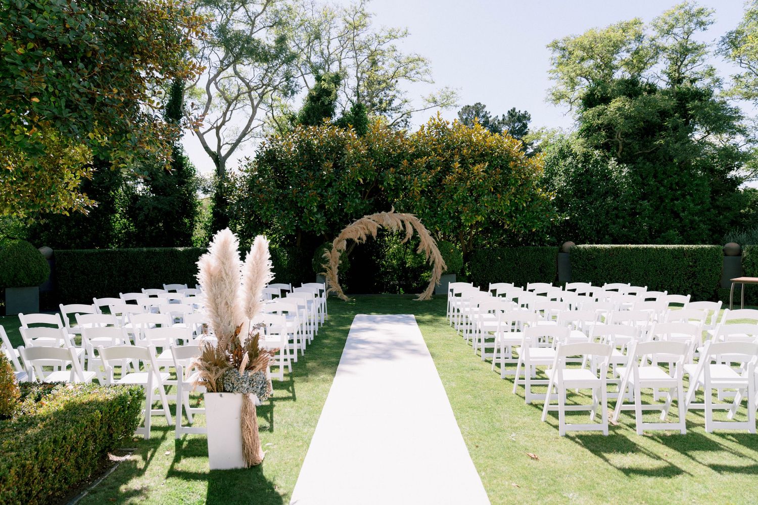 White wooden chairs set up in rows outdoors under green trees for a garden wedding ceremony.