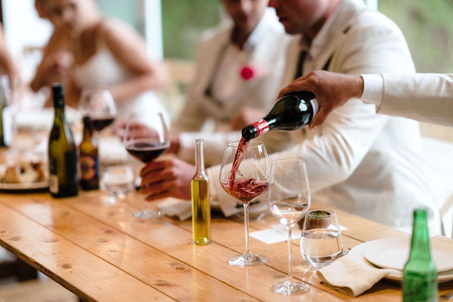 Someone pouring red wine into glasses at a wooden dining table.