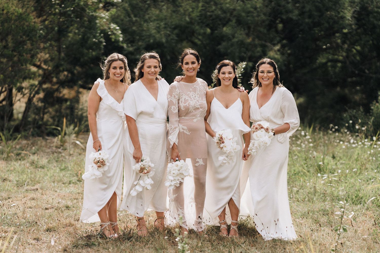 Group of bridesmaids in flowing white dresses pose together on a grassy hillside.