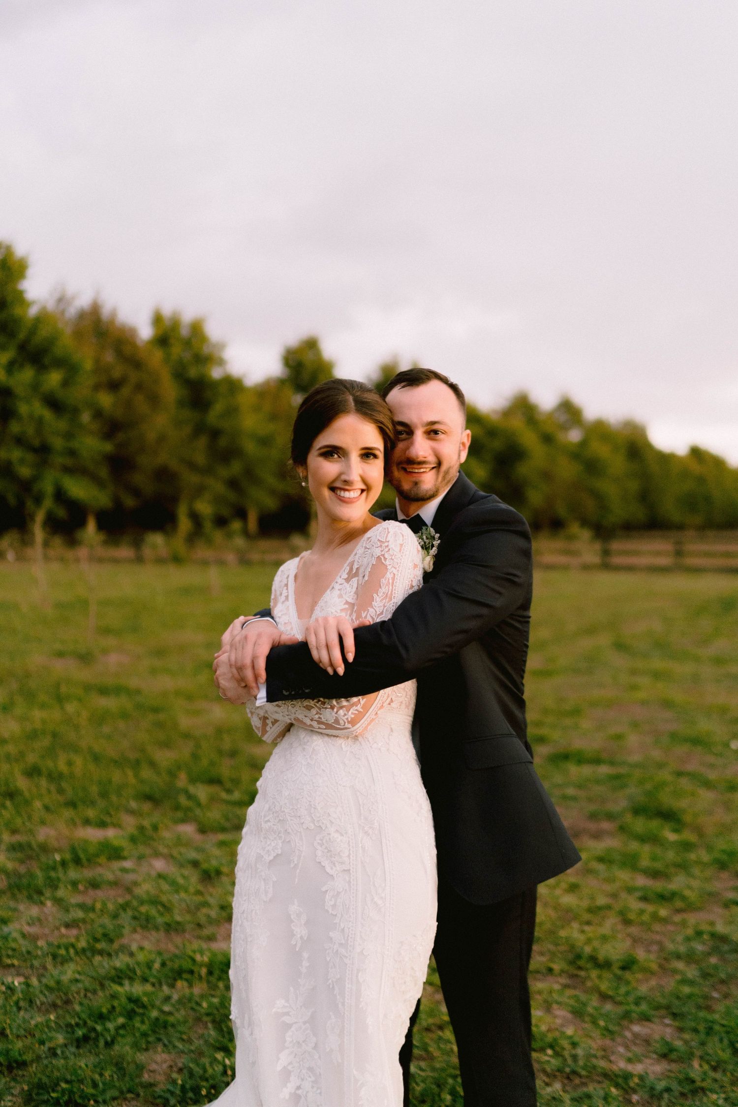 A couple embraces in formal wedding attire while standing in a sunlit field.