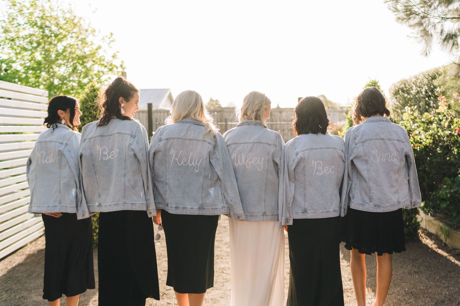 A wedding party shows off matching denim jackets from behind in a garden setting.