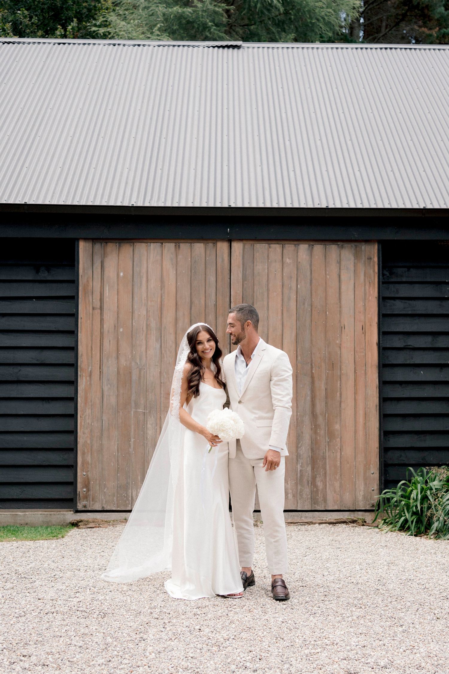 Newlywed couple in white attire pose in front of rustic black barn with wooden door and metal roof.