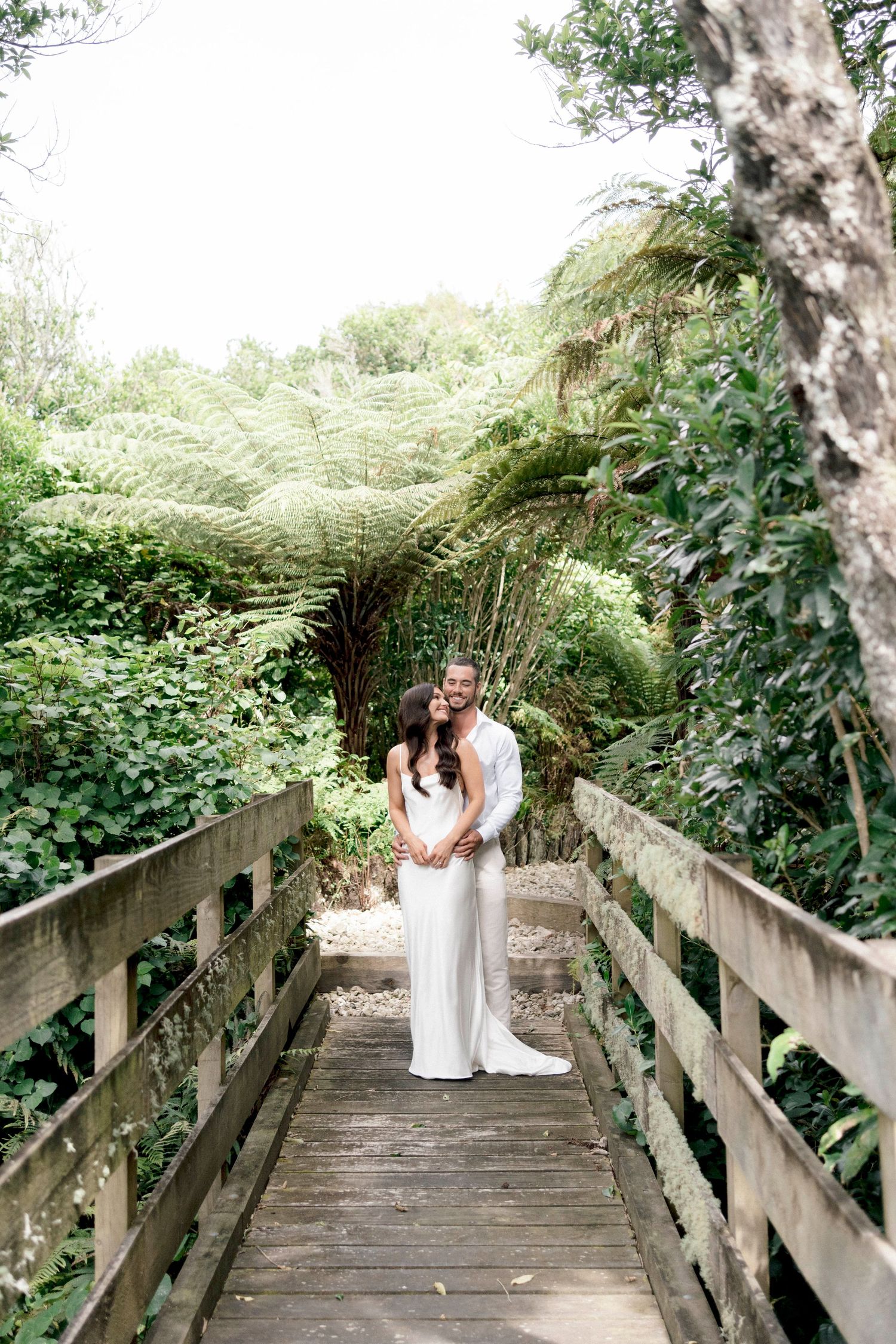 A couple embraces on a wooden boardwalk surrounded by lush ferns and greenery in a romantic outdoor setting.