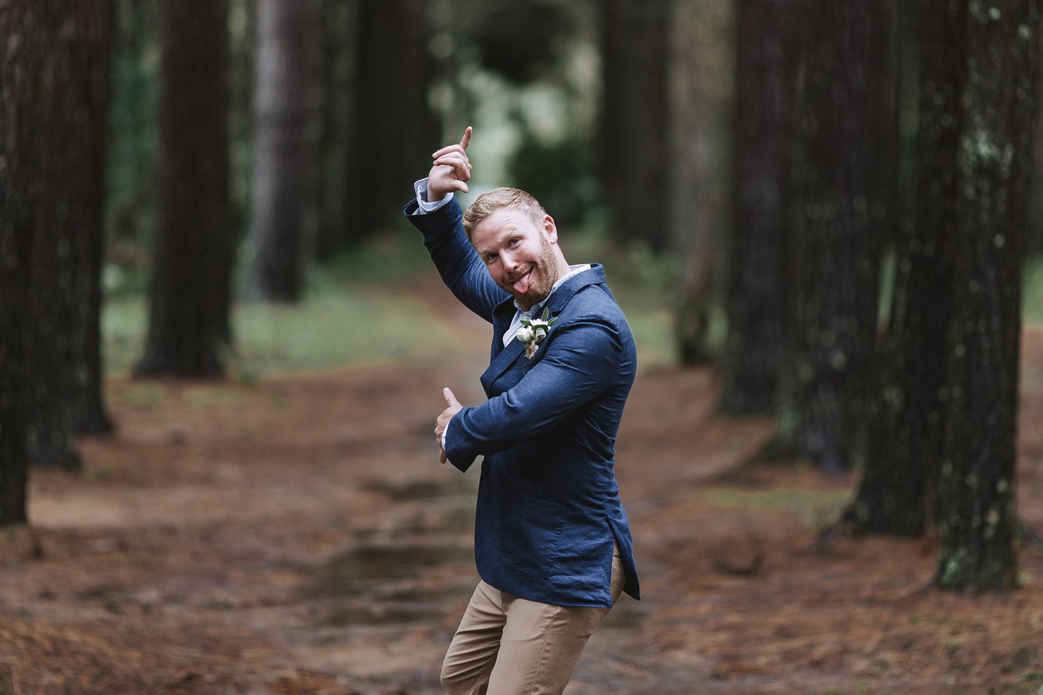 A person dances joyfully on a path through a pine forest while wearing a navy blazer and khaki pants.