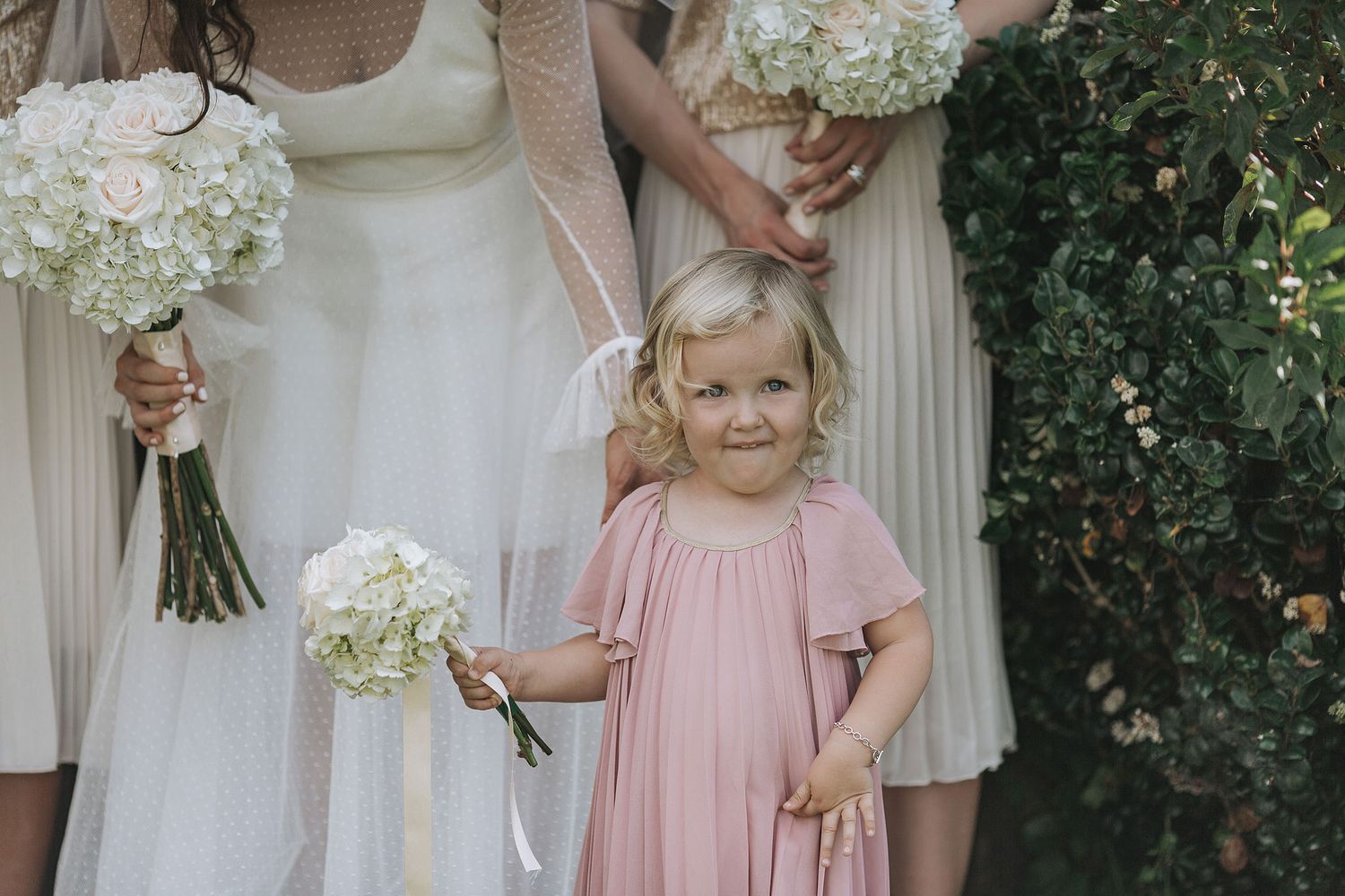 A small child in a pink dress holds a white floral bouquet while standing next to people in light-colored dresses.
