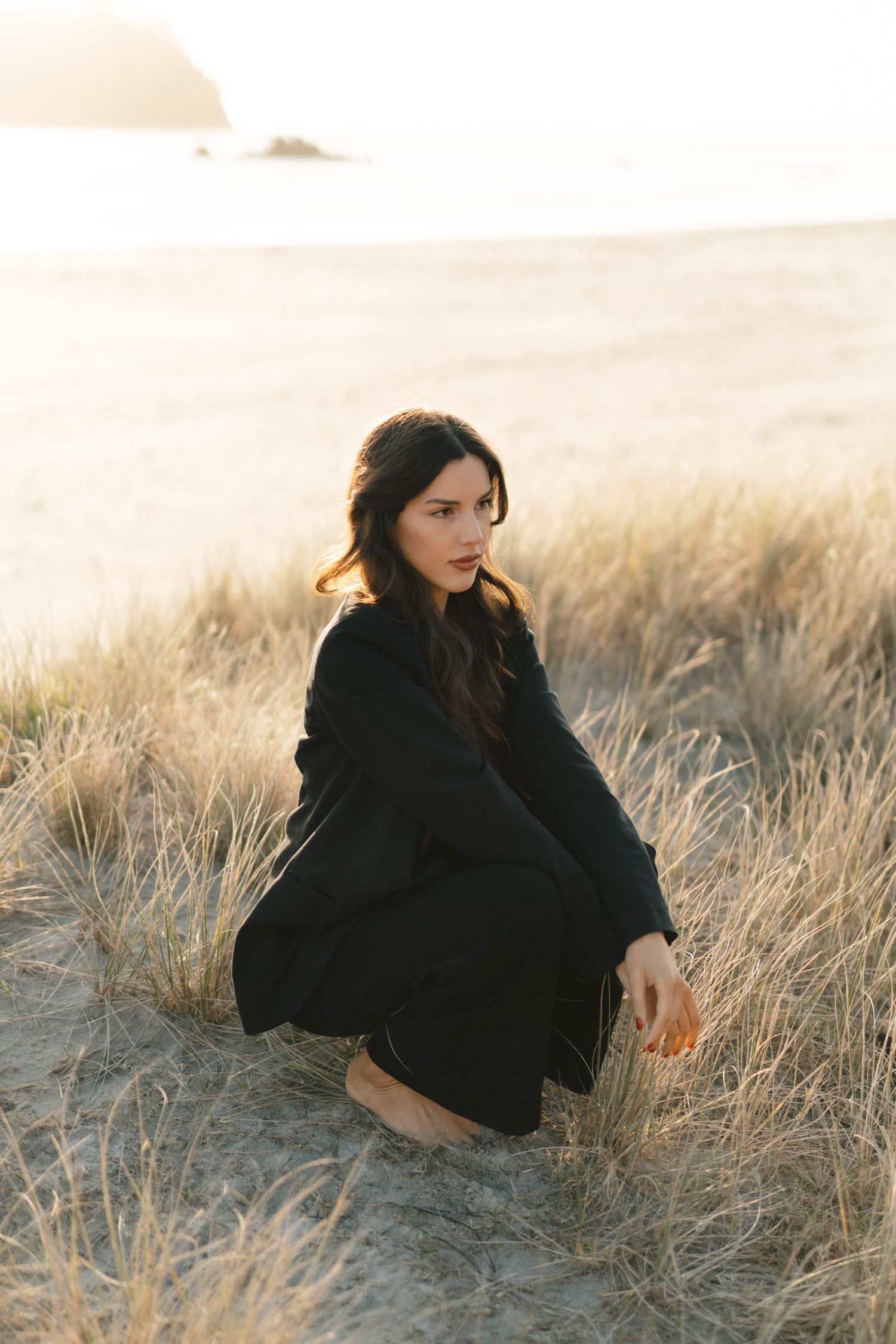 Silhouette crouches in tall beach grass during sunset with ocean view.