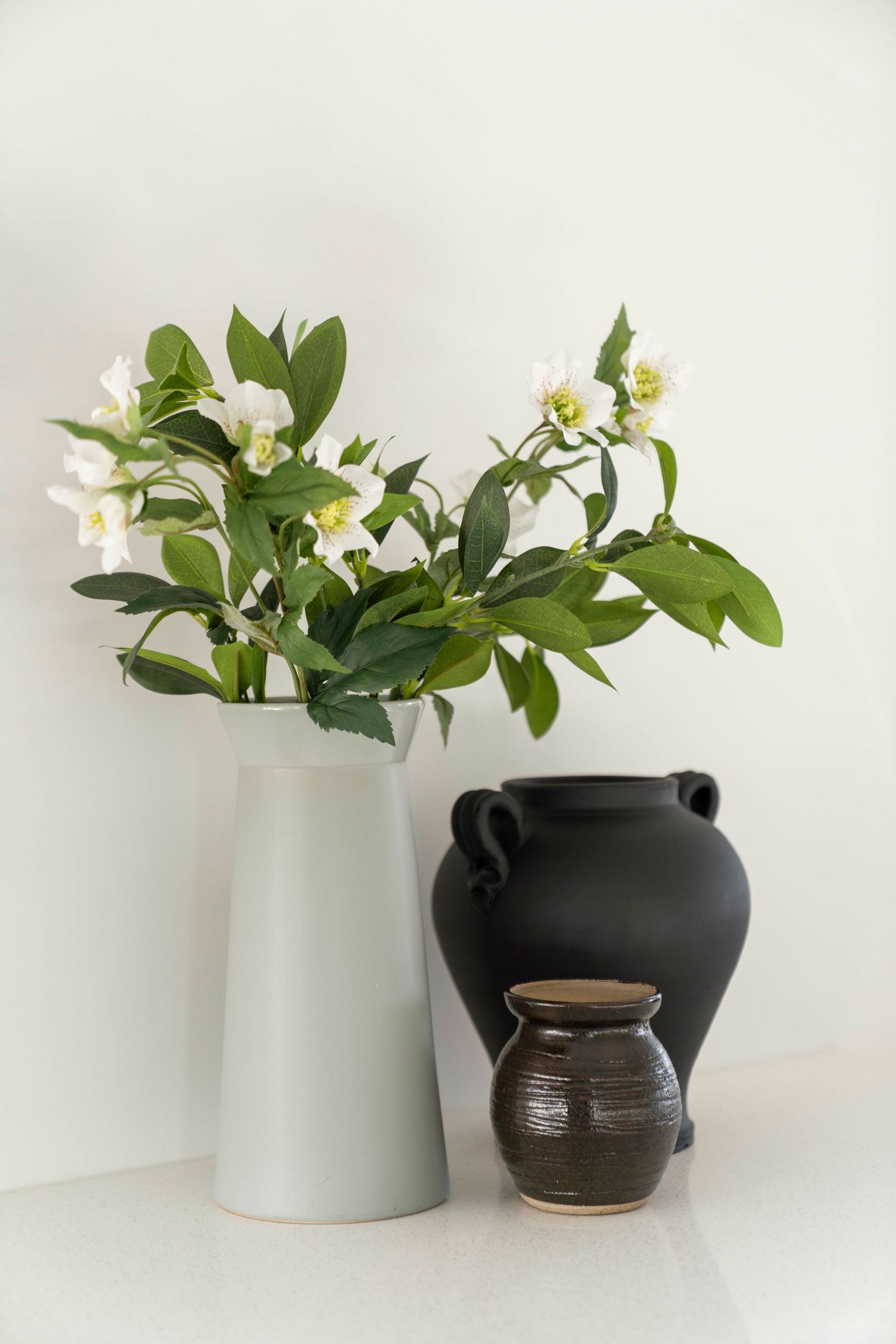 Elegant arrangement of white flowers in a tall vase alongside black and brown ceramic pots.