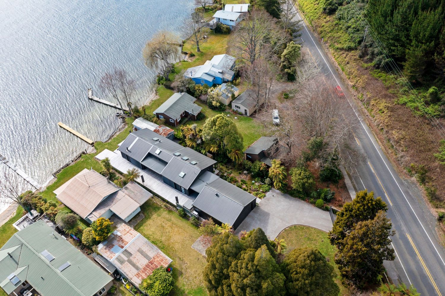 Aerial drone view of waterfront houses and properties along a coastal road with evergreen trees.