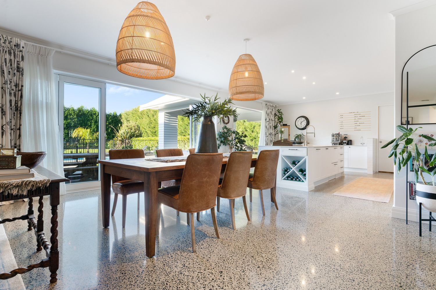 Elegant dining area with wooden table, woven pendant lights, and polished concrete floors connecting to a modern kitchen.