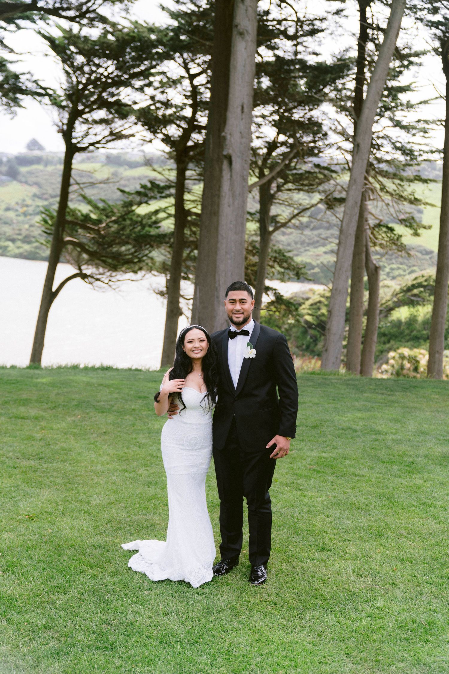 A newlywed couple poses for romantic photos on a grassy hillside with pine trees and water in the background.