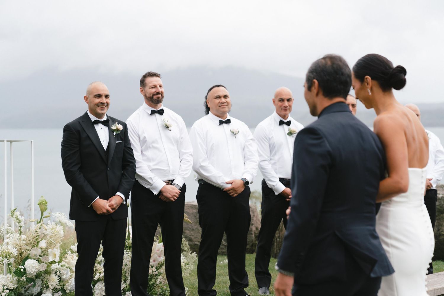 Wedding party stands at an outdoor ceremony overlooking a cloudy landscape with white floral arrangements.
