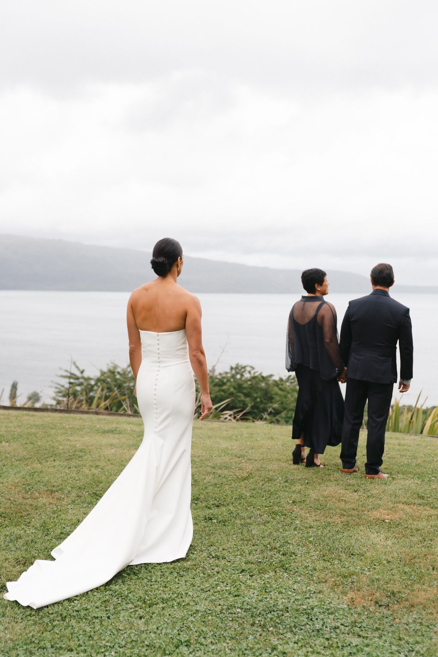 A bride in a white dress stands on a grassy hill overlooking a lake while others walk ahead.