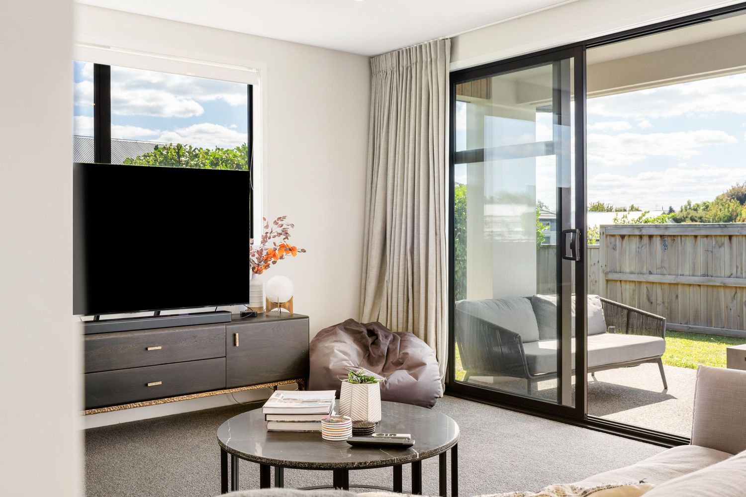 Modern living room with TV mounted on dark cabinet, sliding glass doors, and round coffee table in natural light.