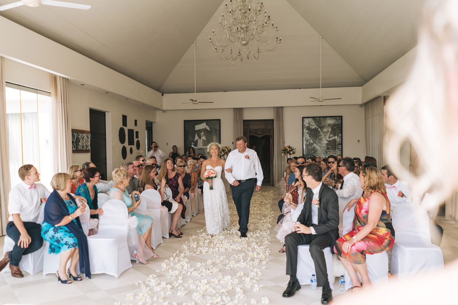 Wedding ceremony in an elegant white room with guests seated in rows and white petals lining the aisle.