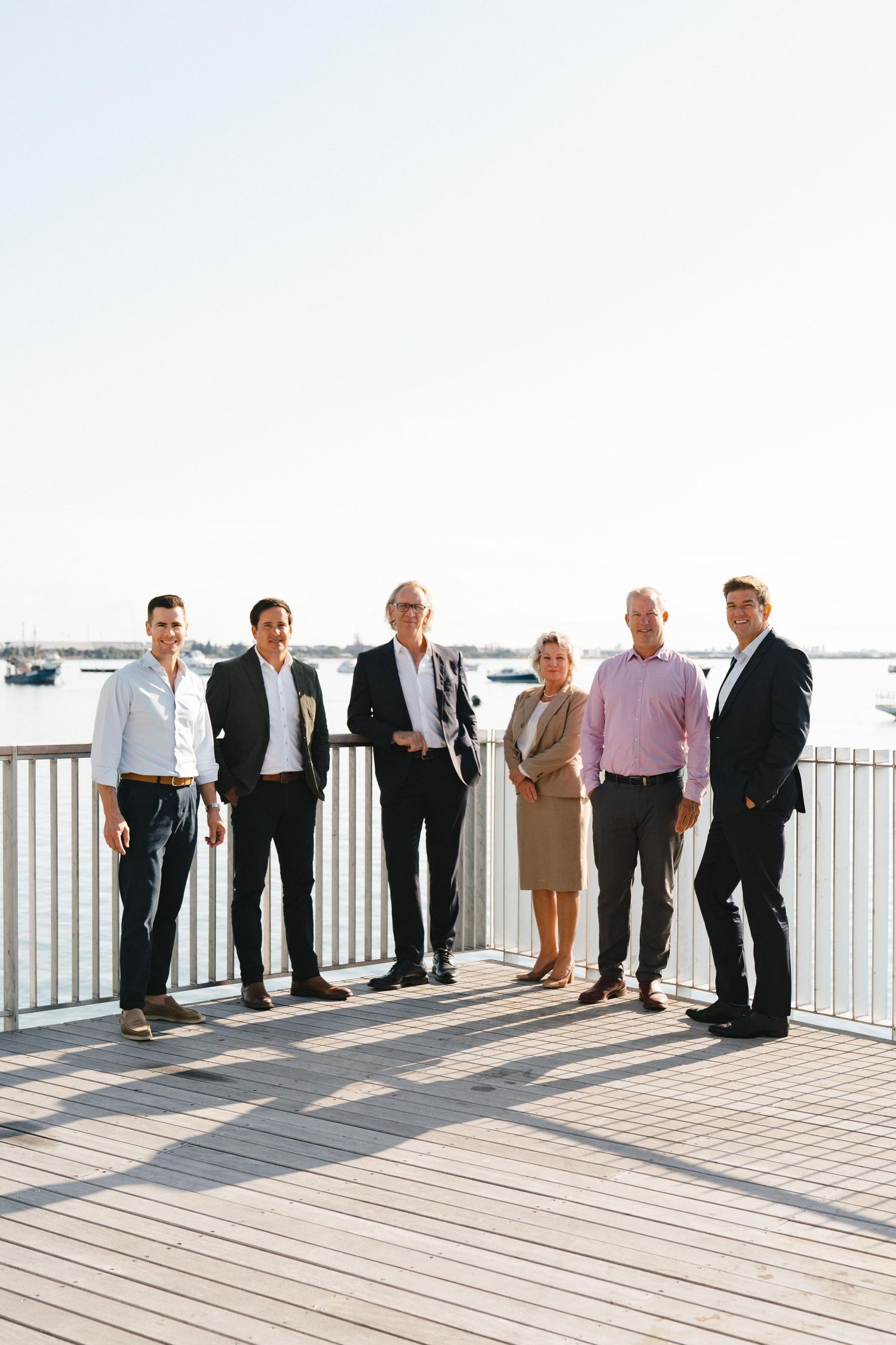 Business team standing on marina boardwalk wearing formal attire with boats and waterfront in background.