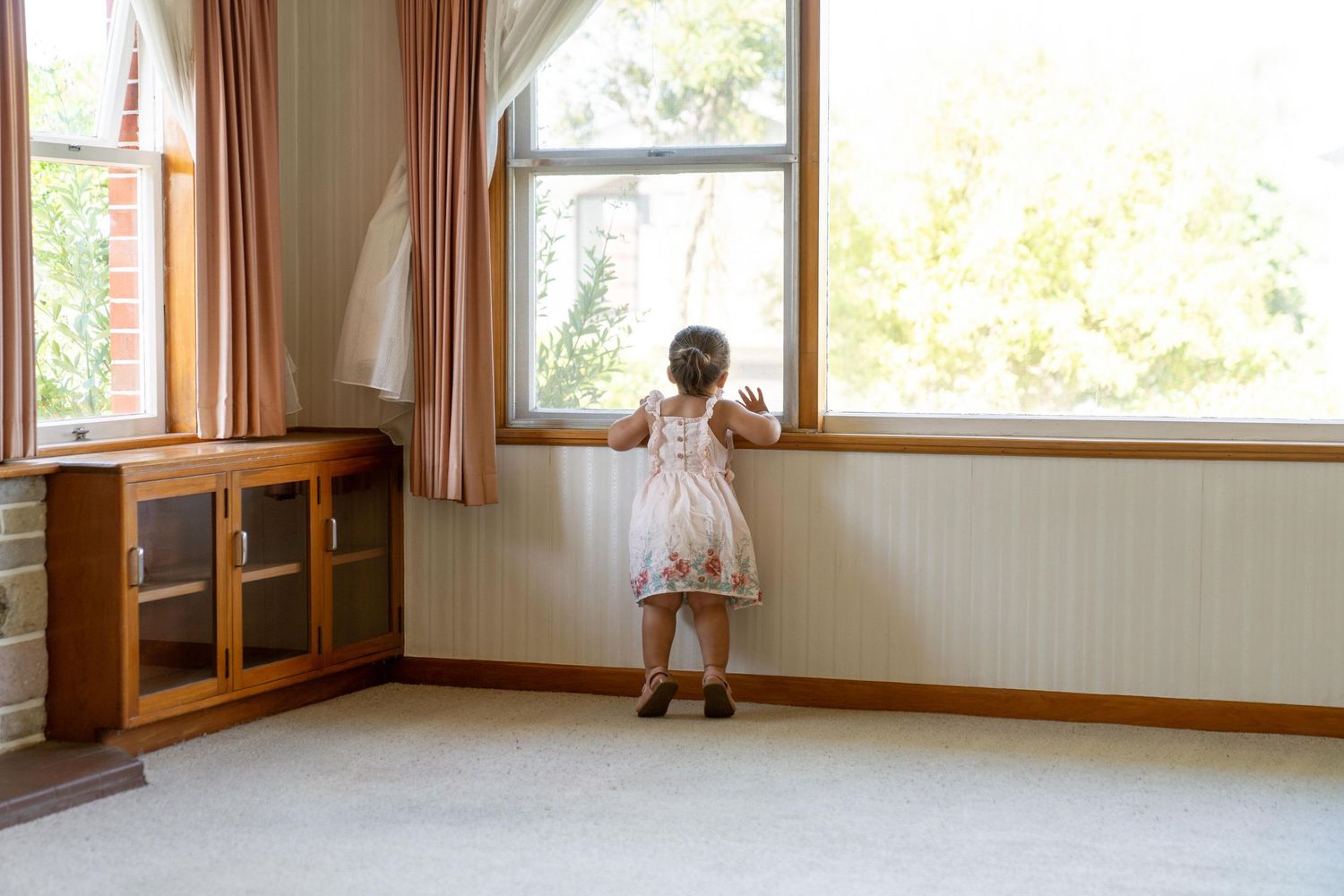 Small child looking out large windows in a room with wooden furniture and cream walls.